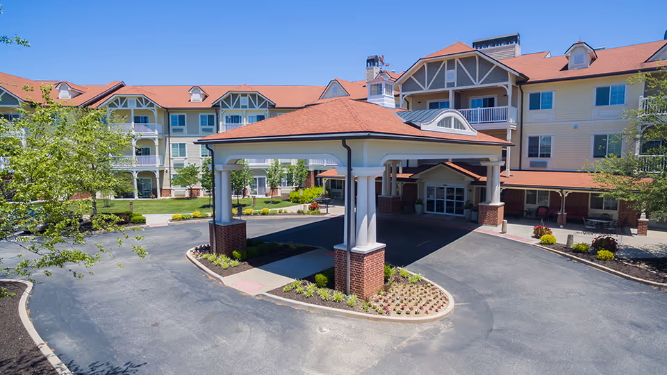 Front exterior view of Crestview Senior Living facility showing a large covered entrance with brick pillars, a circular driveway, landscaped flower beds, and a multi-story building with balconies and a red roof under a clear blue sky.