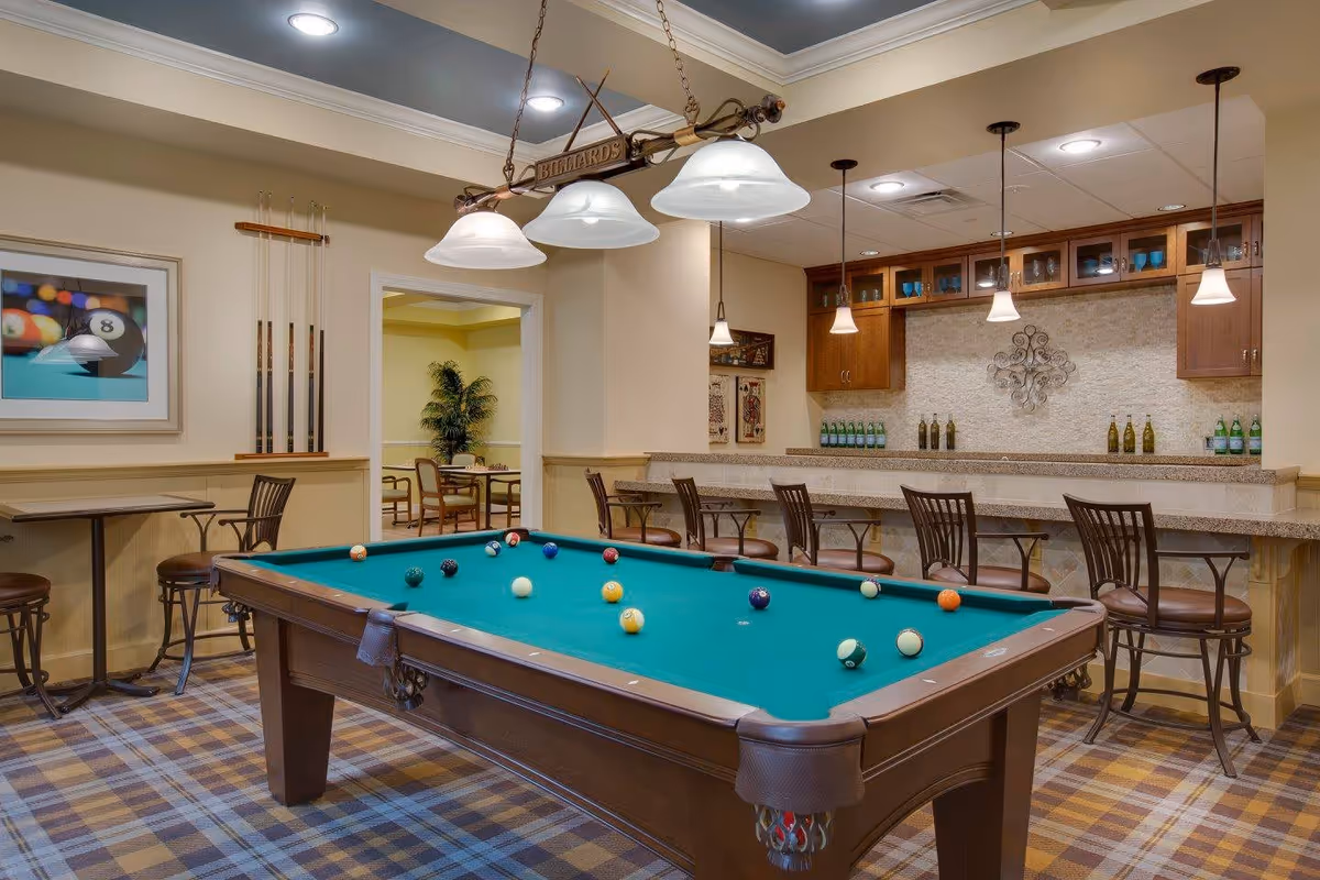 A recreational room featuring a billiards table with balls arranged on the green felt surface. Above the table hangs a light fixture with three lamps and a sign that reads 'BILLIARDS'. To the right, there is a bar area with high chairs, pendant lights, and shelves with bottles and glassware. On the left wall, there is a framed picture of billiard balls and a rack holding pool cues. In the background, another room with tables and chairs and a potted plant is visible.
