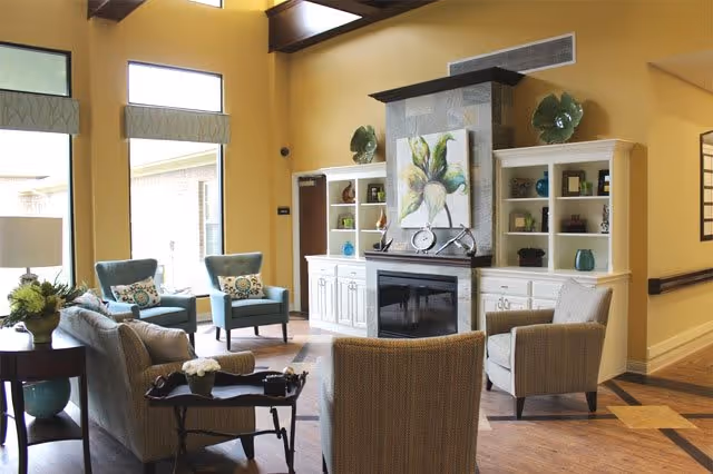 A cozy living room area with a fireplace in the center, flanked by white built-in shelves decorated with vases and ornaments. There are several upholstered chairs and a sofa arranged around a small coffee table. Large windows allow natural light to fill the room, and the walls are painted a warm yellow color.