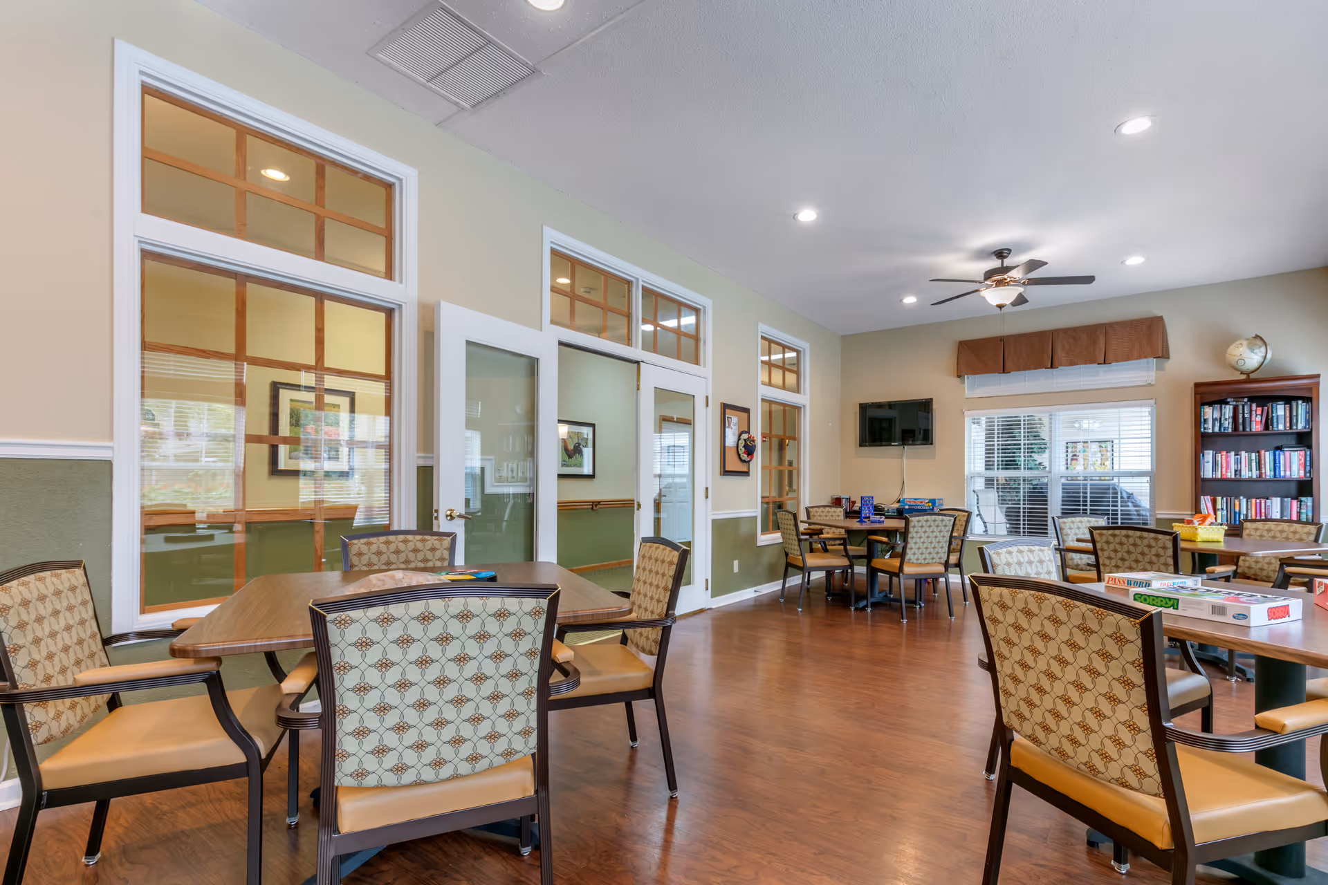 A bright and spacious common area with multiple tables and chairs arranged for group activities. The room features large windows with white blinds, a ceiling fan with lights, a wall-mounted TV, and a bookshelf filled with books and board games. The floor is wooden, and the walls are painted in light colors with white trim.