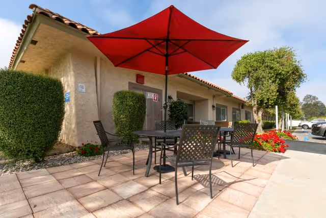 Outdoor patio area at Camarillo Healthcare Center with a round metal table and four matching chairs under a large red umbrella. The patio is paved with tiles and surrounded by neatly trimmed bushes and red flowers. The building with a tiled roof and beige walls is visible in the background along with a parking area and trees.