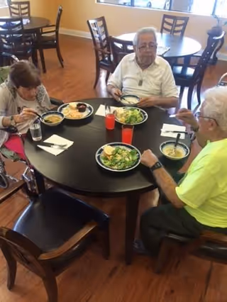 Three elderly people sitting around a round table in a dining area, eating a meal that includes soup and salad. The room has wooden floors and several other tables and chairs in the background.