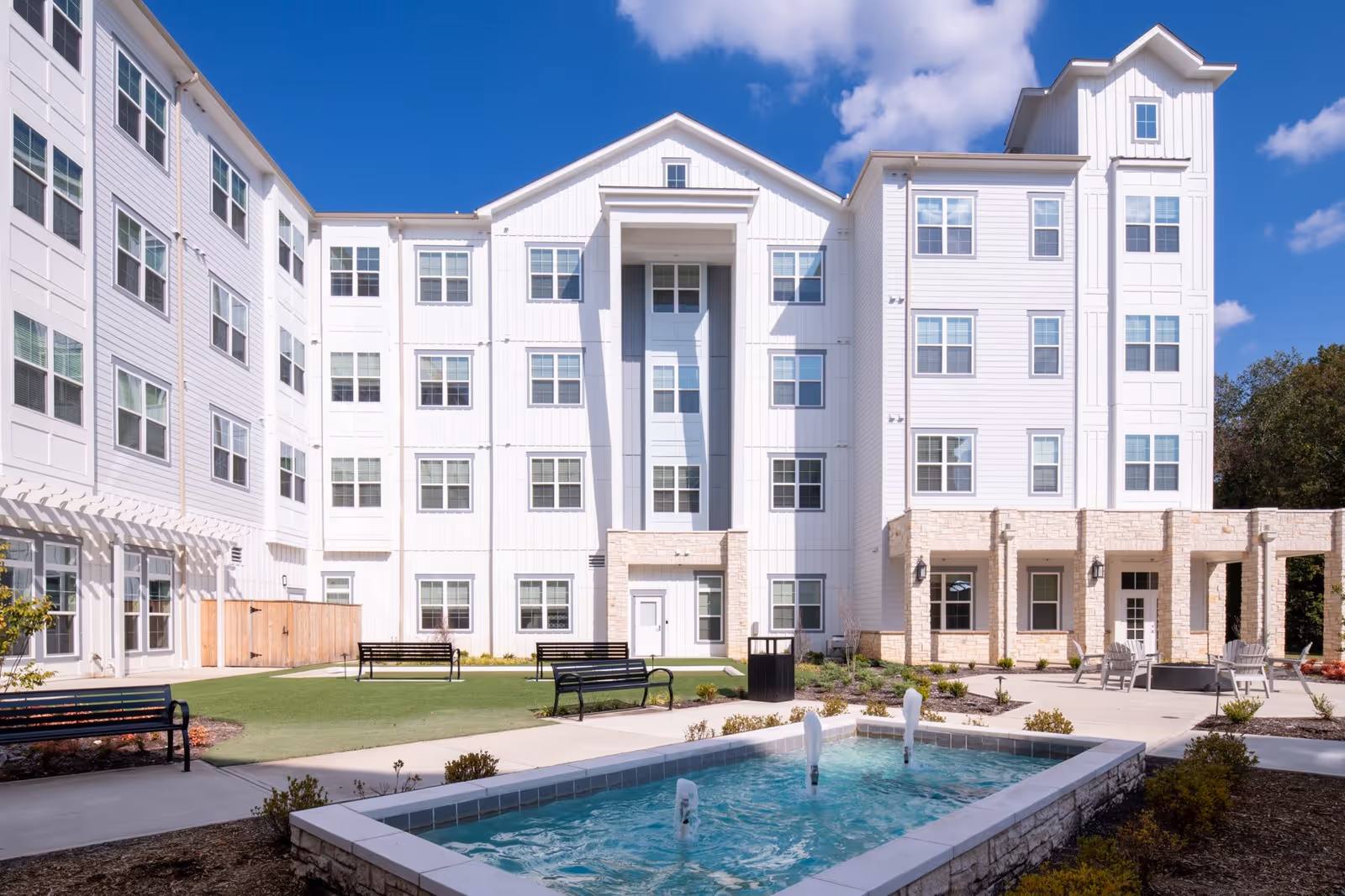 Outdoor courtyard area of The Summit of Germantown senior living facility featuring a rectangular water fountain with three water jets, several black benches, a well-maintained lawn, and a multi-story white building with many windows under a blue sky with some clouds.