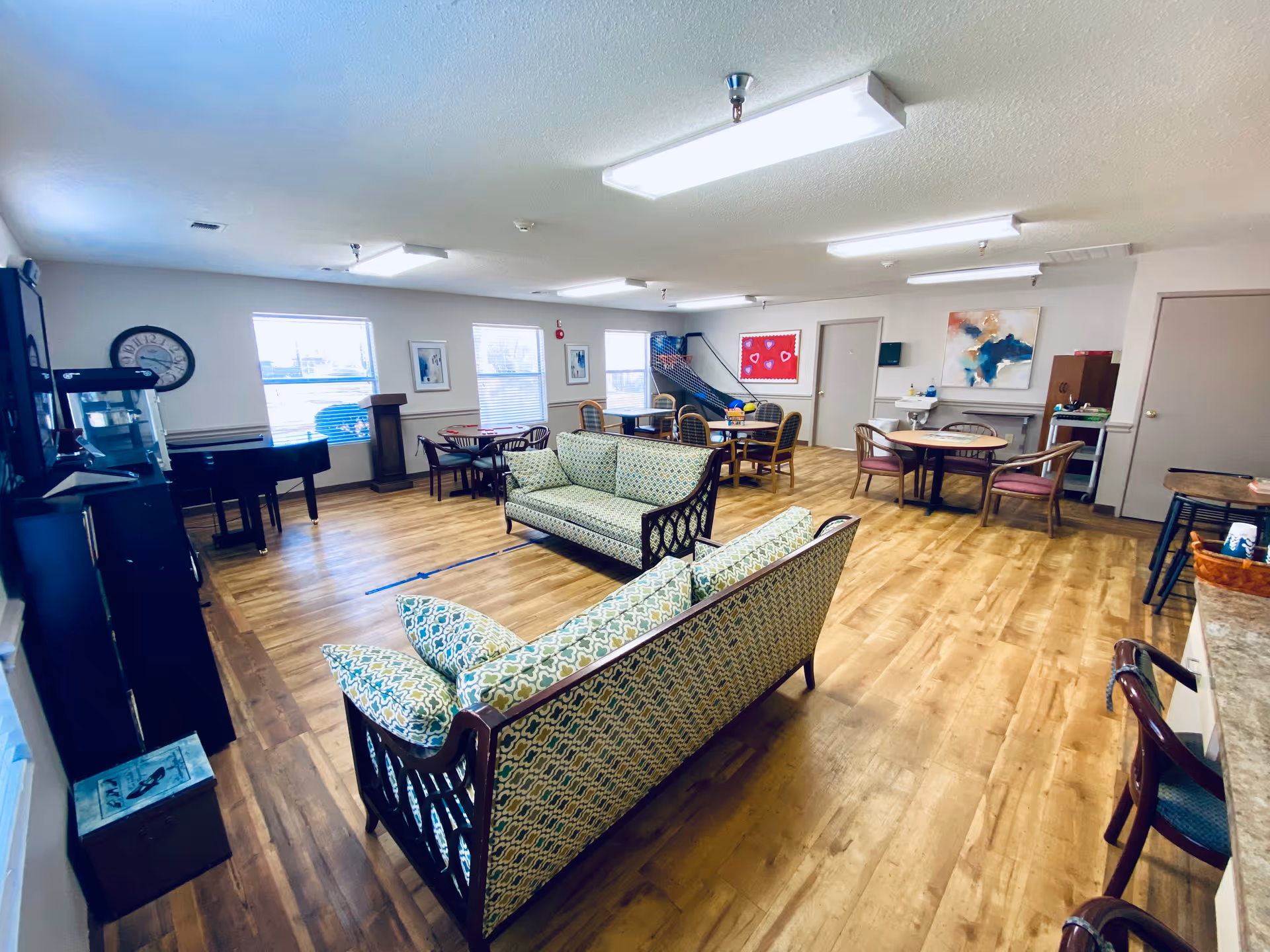 A spacious common area in Mullican Care Center with wooden flooring, two patterned sofas facing each other, multiple round tables with chairs, a piano near the windows, and a basketball arcade game in the corner. The room is well-lit with ceiling lights and has various decorations on the walls.