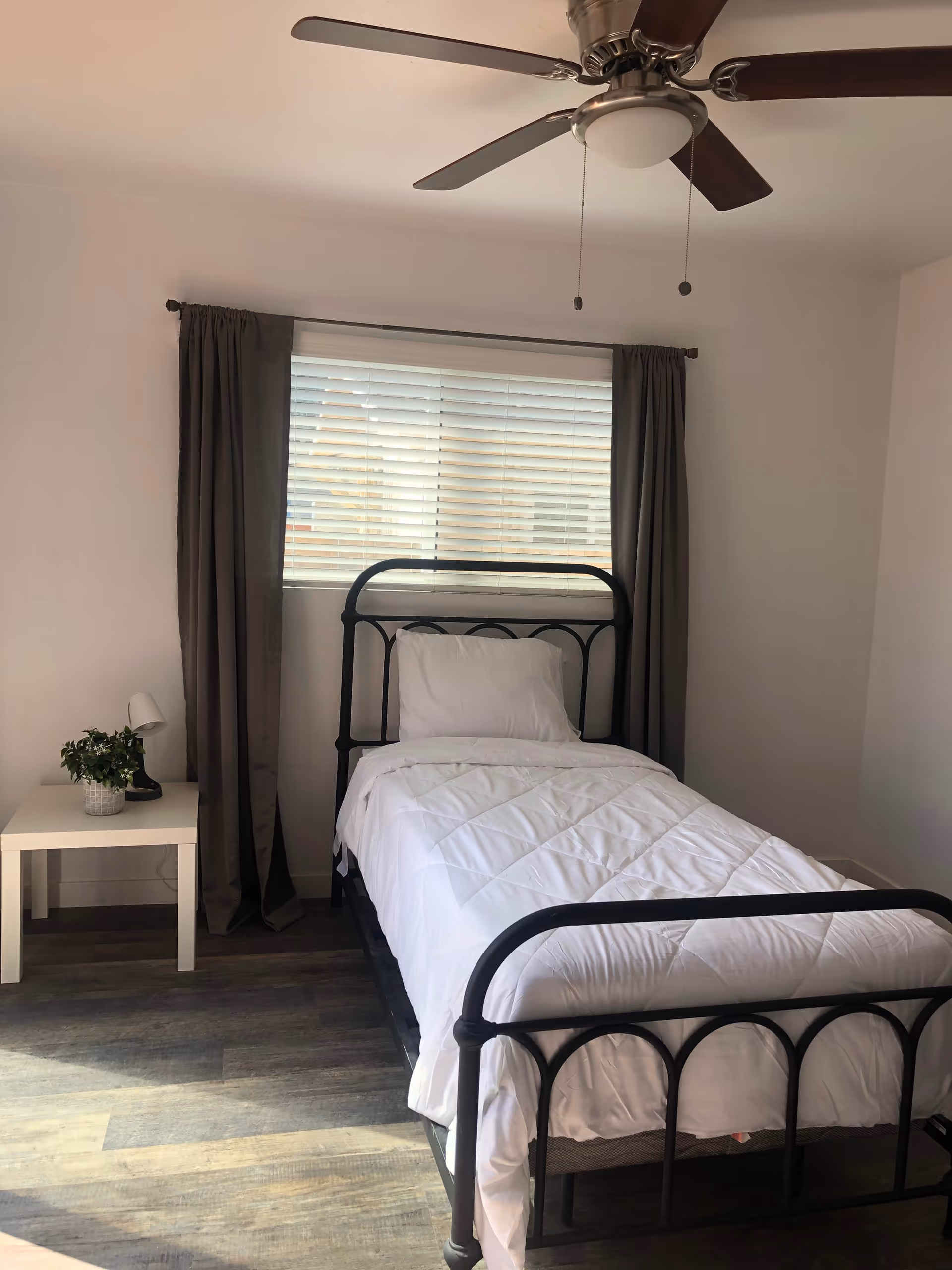 A small bedroom with a single black metal bed frame, white bedding, and a pillow. There is a window with white blinds and dark curtains behind the bed. A white side table with a small plant and a lamp is placed next to the bed. A ceiling fan with wooden blades is visible above.