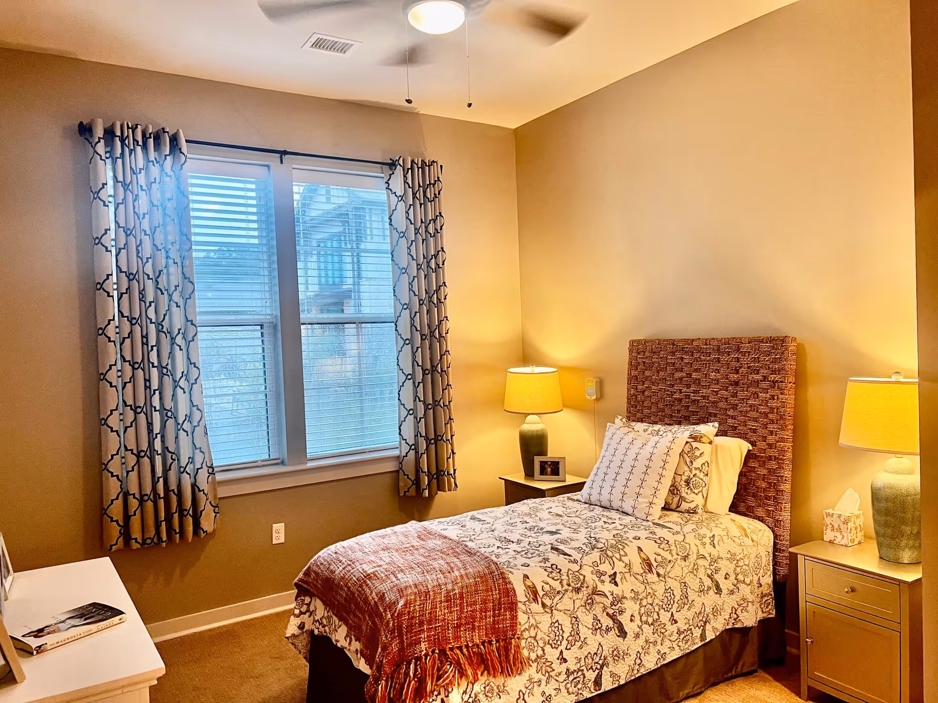 Well-lit bedroom with a single bed, patterned curtains over a double window, bedside tables with lamps, and warm beige walls.