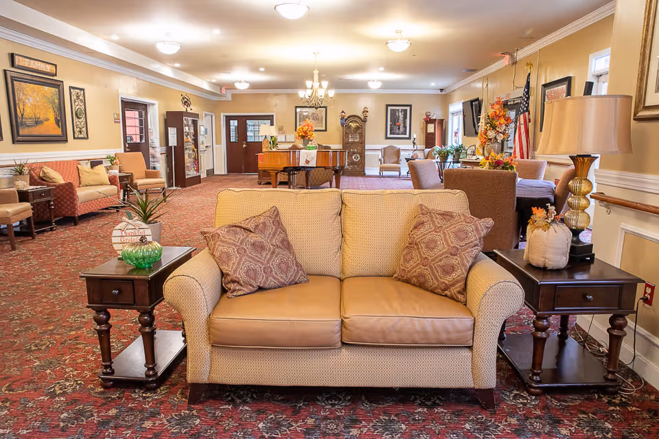 A cozy nursing home common area with a beige loveseat featuring two patterned pillows in the foreground. On either side of the loveseat are dark wooden side tables, one with a green glass bowl and a small plant, the other with a lamp and a decorative pumpkin. The room has a red patterned carpet, beige walls, and various chairs and tables arranged throughout. There are framed pictures on the walls, an American flag, and a piano in the background.
