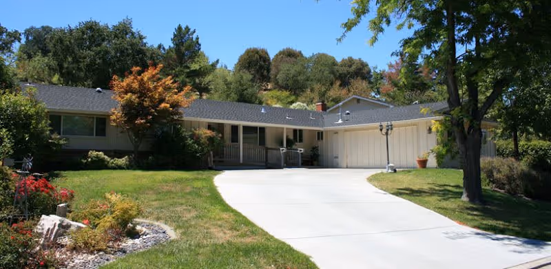 Single-story residential building with a wide driveway, surrounded by green lawn, trees, and shrubs under a clear blue sky.