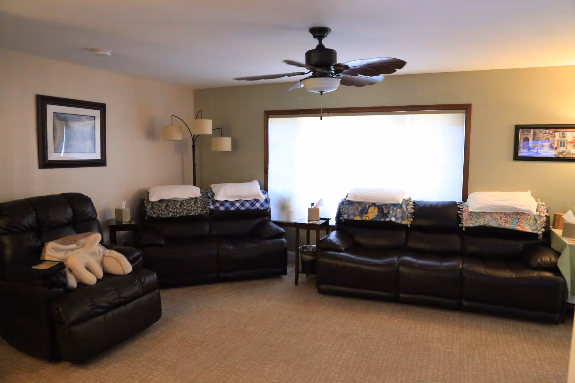 A cozy living room with two black leather sofas and one black leather recliner chair. Each sofa has folded blankets and white towels placed on the backrests. There is a floor lamp with three lights in the corner, a ceiling fan with leaf-shaped blades, and framed artwork on the walls. A large window with blinds lets in natural light.