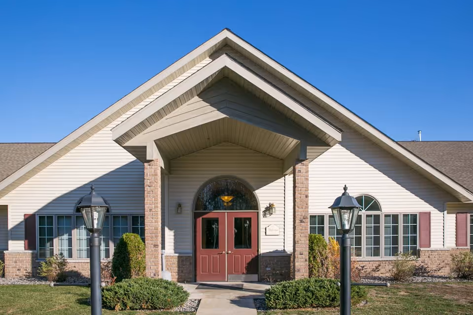 Front exterior view of a single-story assisted living facility building with beige siding, brick accents, a peaked roof, red double doors, and two black lamp posts along the walkway leading to the entrance under a clear blue sky.