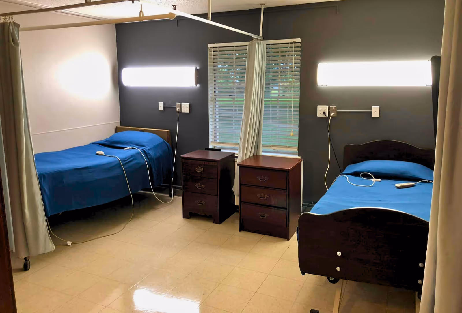 A room with two hospital-style beds separated by a window with blinds and two wooden nightstands. Each bed has a blue blanket and a call button remote. The walls are painted dark gray and white, and there are bright wall-mounted lights above each bed. Privacy curtains are partially visible on both sides of the room.