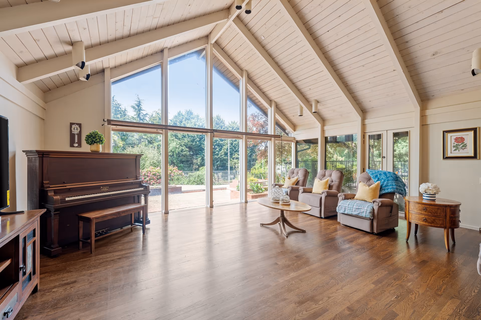 A spacious living room with high vaulted ceilings and large floor-to-ceiling windows offering a view of a garden outside. The room features a dark wooden piano with a matching bench, three cushioned armchairs with yellow pillows and blue throws, a round wooden coffee table, a small wooden side table with a vase of flowers, and a framed flower artwork on the wall.
