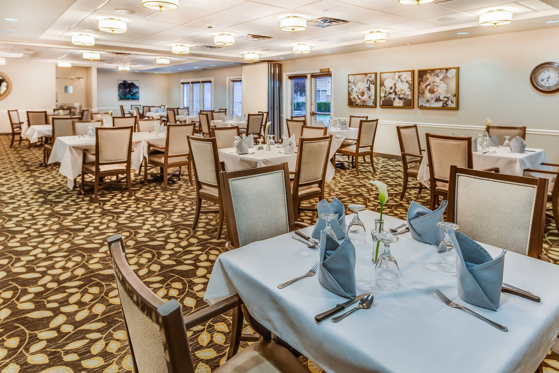 A spacious dining room with multiple tables covered in white tablecloths, each set with blue folded napkins, glassware, and silverware. The room has patterned carpet flooring, beige walls adorned with floral artwork, and ceiling lights providing warm illumination.