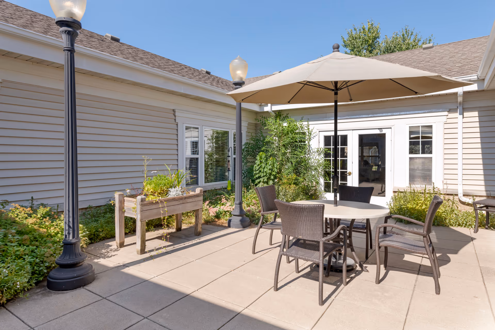 Outdoor patio area with a round table, four wicker chairs, and a large beige umbrella. The patio is surrounded by beige siding walls with windows and a glass door. There are two black lamp posts and various green plants and shrubs around the patio.