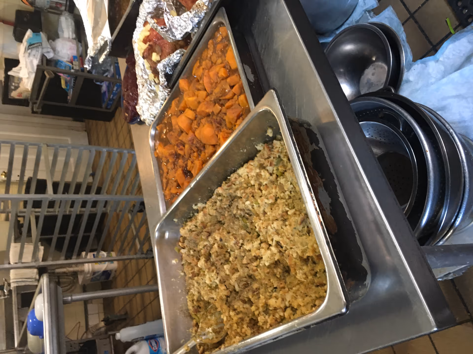 Metal food pans containing stuffing and sweet potatoes on a stainless-steel serving cart in a commercial kitchen.