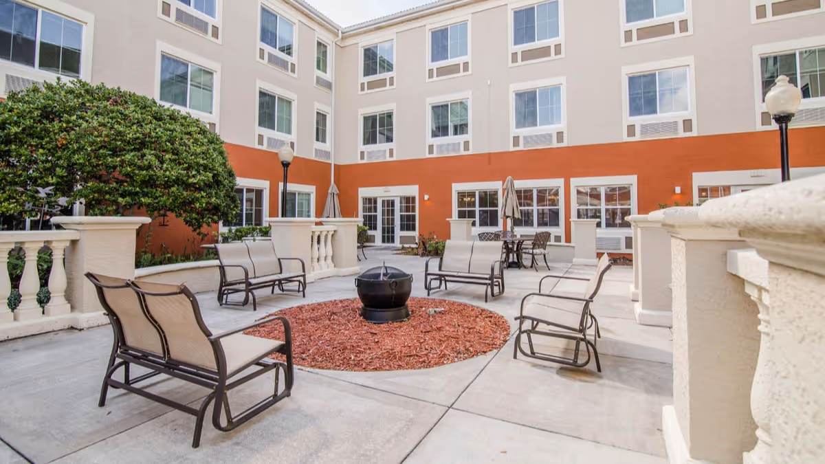 Outdoor courtyard area at Brookdale Lake Orienta with multiple metal and mesh chairs arranged around a circular fire pit filled with red mulch. The courtyard is surrounded by a three-story building with large windows and beige and orange walls. There are also some tables with umbrellas and decorative lamp posts.