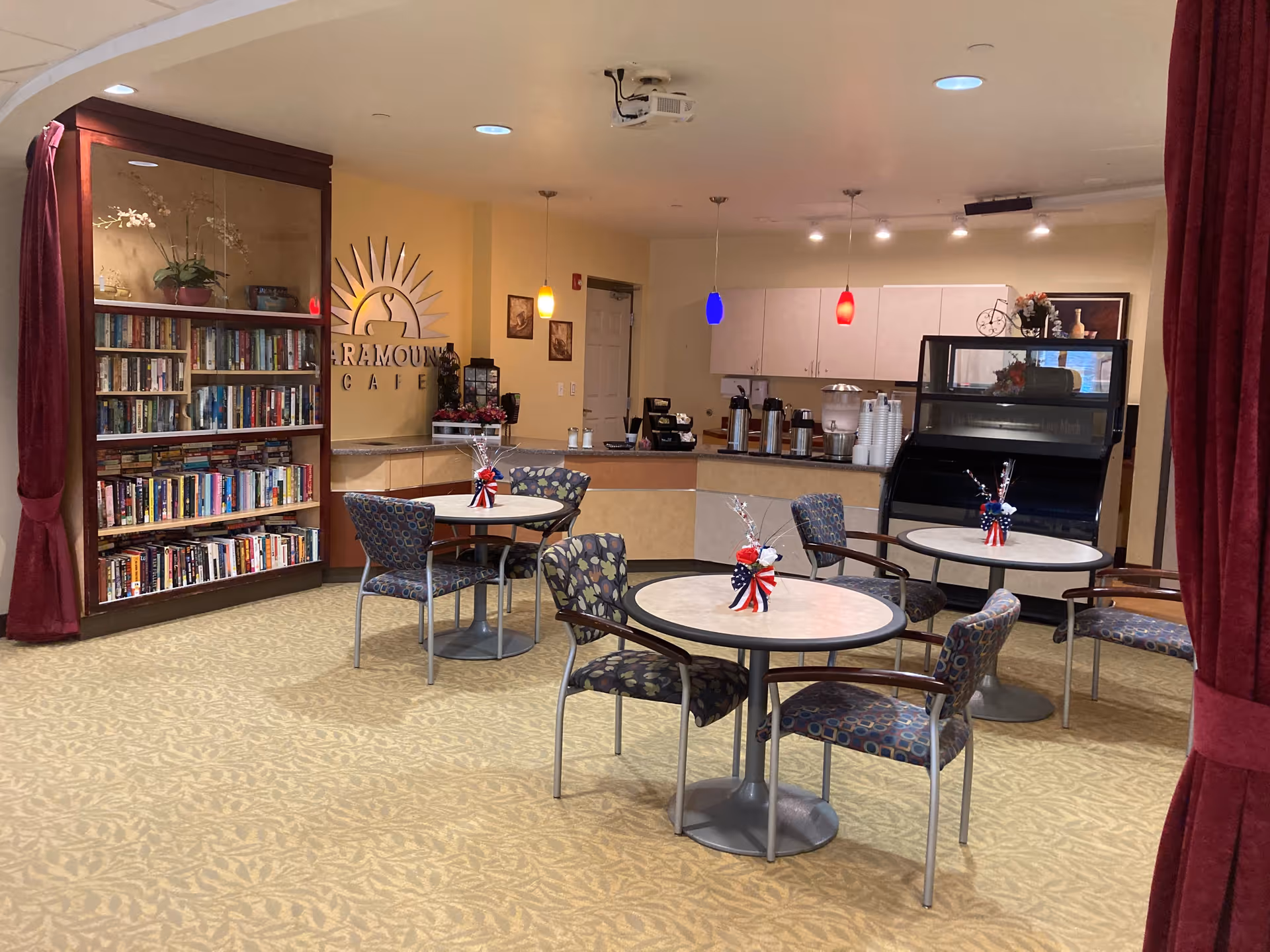 Interior view of a senior living facility cafe area with round tables and chairs arranged for seating. Each table has a small patriotic-themed centerpiece. There is a bookshelf filled with books on the left side, and a counter with coffee dispensers and cups in the background. Pendant lights hang from the ceiling, and a sign on the wall reads 'Paramount Cafe'.