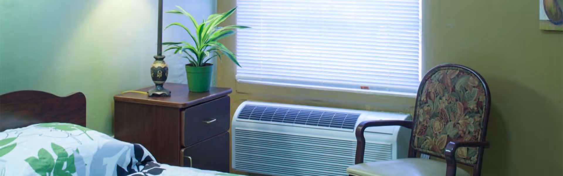 A small bedroom with a single bed covered in a white and green patterned blanket, a wooden nightstand with a lamp and a potted plant, a window with closed blinds, an air conditioning unit below the window, and a floral upholstered armchair.