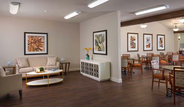 A bright communal living area with sofas, a coffee table, framed artwork, and dining tables and chairs in the background.