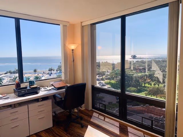 A bright office corner with a desk, office chair, printer, and laptop positioned next to large windows offering a scenic view of the ocean, palm trees, and a park area under a clear blue sky.