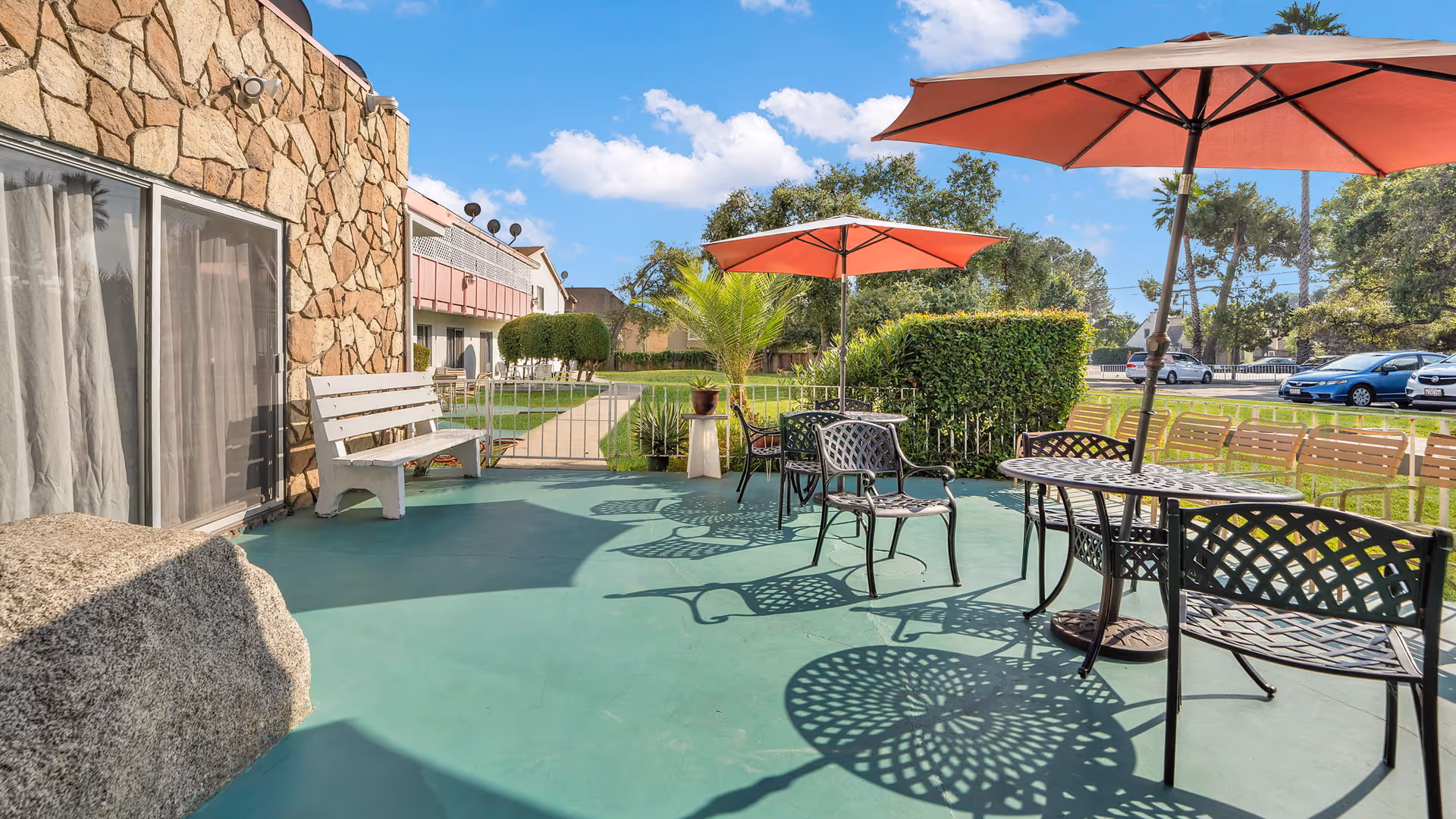 Outdoor patio area at Glen Park at Monrovia with metal tables and chairs under large umbrellas, a white bench against a stone wall, green flooring, and a view of a grassy area with trees and parked cars in the background under a blue sky with some clouds.