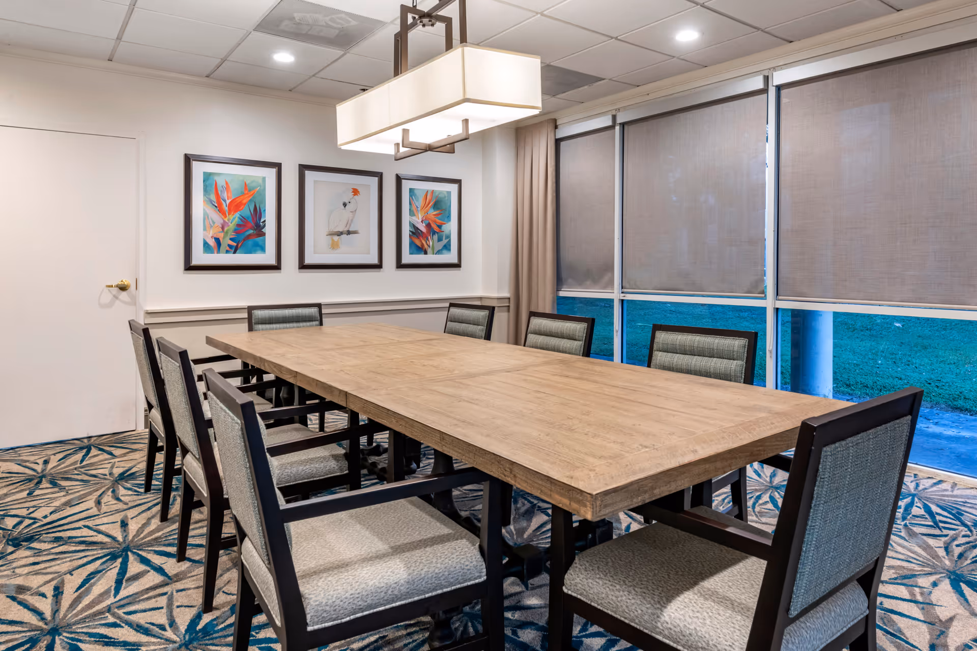 A bright dining/meeting room with a long wooden table surrounded by upholstered chairs, framed artwork on the wall, and large windows with roller shades.