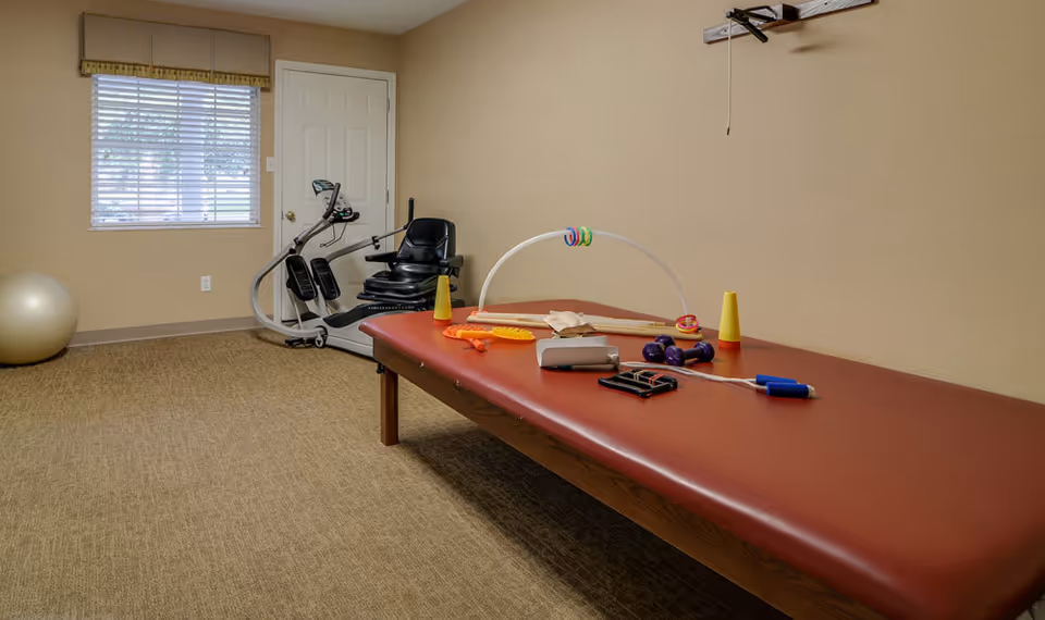 A therapy or exercise room with a padded treatment table covered with various small exercise and therapy equipment, including dumbbells, cones, and rings. In the background, there is a recumbent exercise bike near a window with blinds and a closed door. The walls are beige and the floor is carpeted.