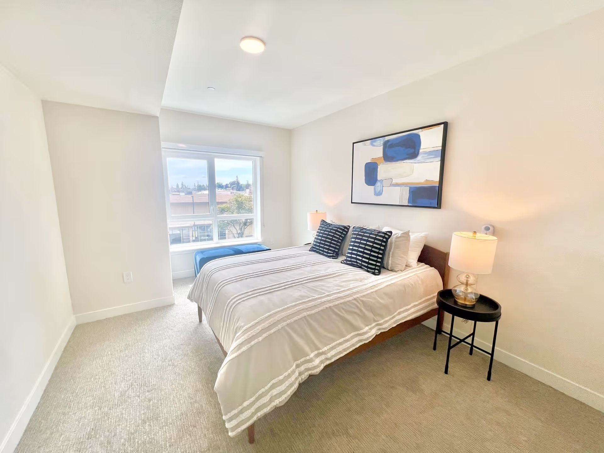 Bright, minimalist bedroom with a made bed, two bedside lamps, abstract art above the headboard, and a large window with a bench.