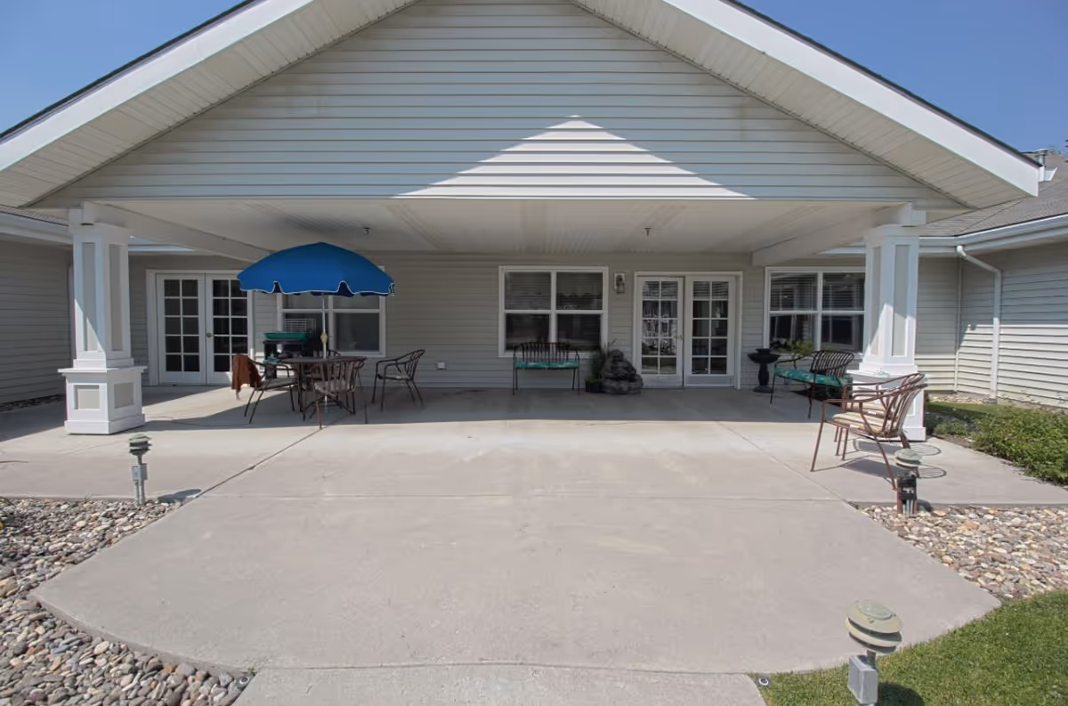 Covered outdoor patio area at a senior living facility with a concrete floor, several metal chairs, a table with a blue umbrella, and two benches. The building exterior is light gray with white trim and multiple glass doors and windows.