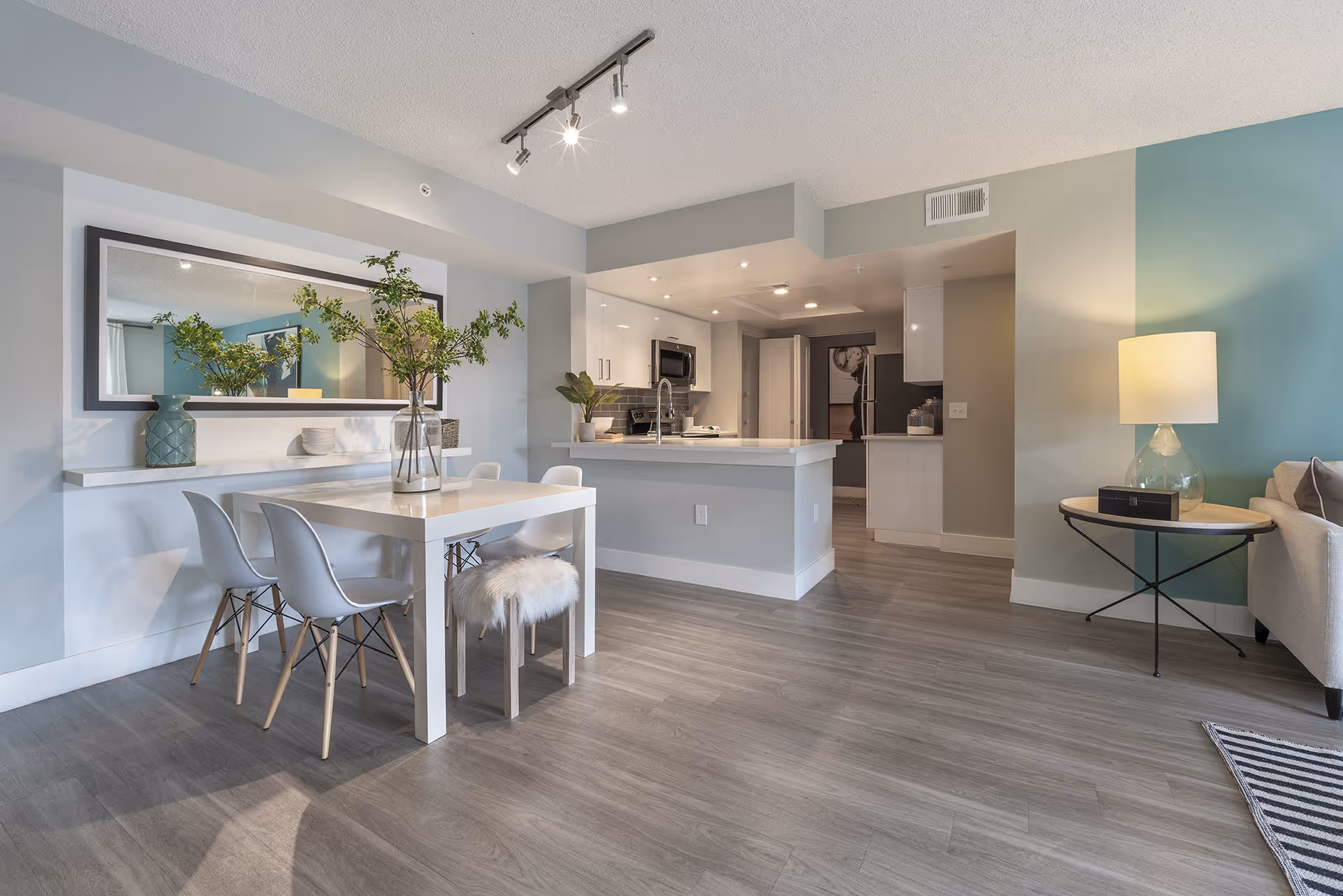 A modern open-plan living space featuring a white dining table with four chairs and a small furry stool, a large mirror on the wall above a floating shelf with decorative vases and greenery, a kitchen area with white cabinets, stainless steel appliances, and a countertop, and a cozy corner with a side table and lamp next to a sofa.