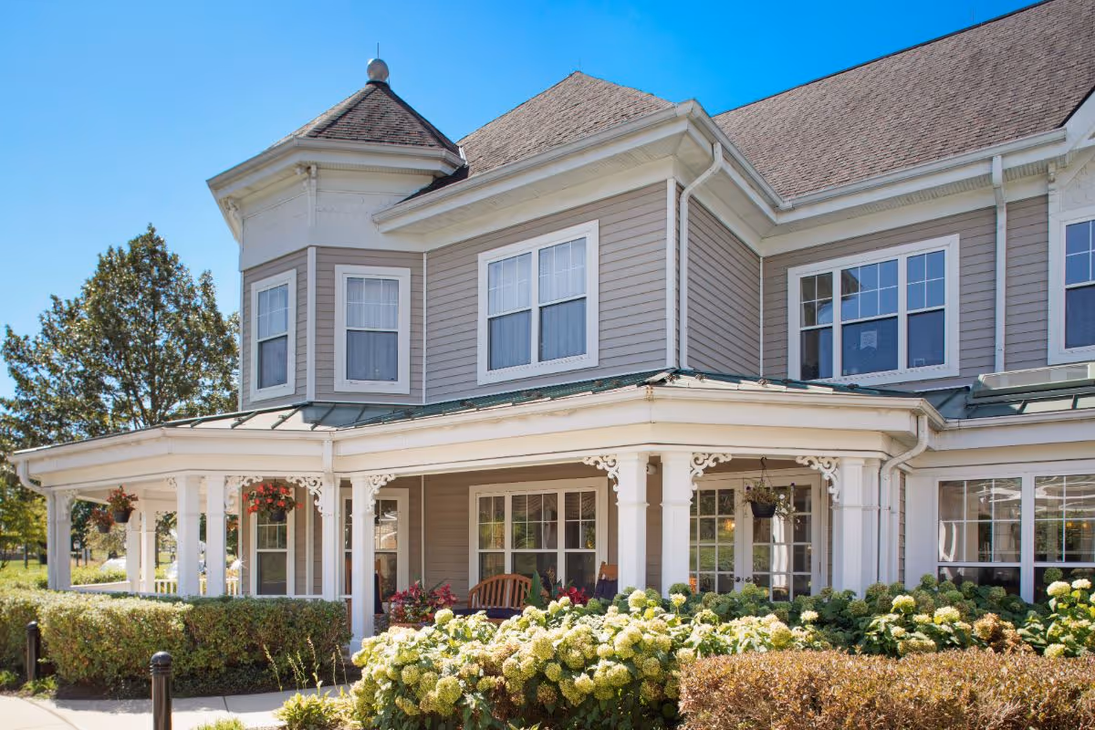 Front exterior of a two-story senior living building with a wraparound porch, decorative columns, and landscaped shrubs and flowers.