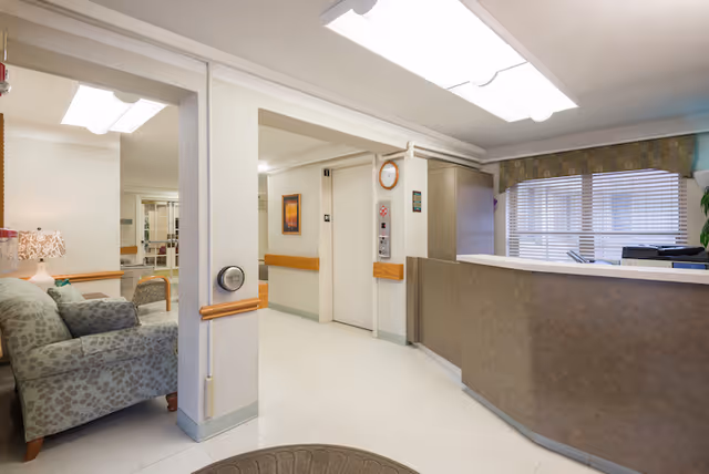 Interior view of a reception area in a healthcare facility showing a front desk with a computer, an elevator with closed doors, a clock on the wall, a patterned armchair with a lamp beside it, and a window with blinds and a valance.