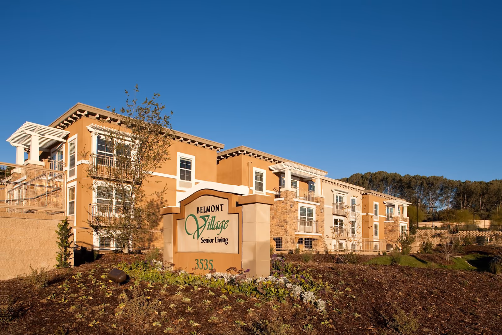 Exterior view of Belmont Village Senior Living Cardiff by the Sea building with clear blue sky, landscaped garden, and a sign displaying the facility name and address.