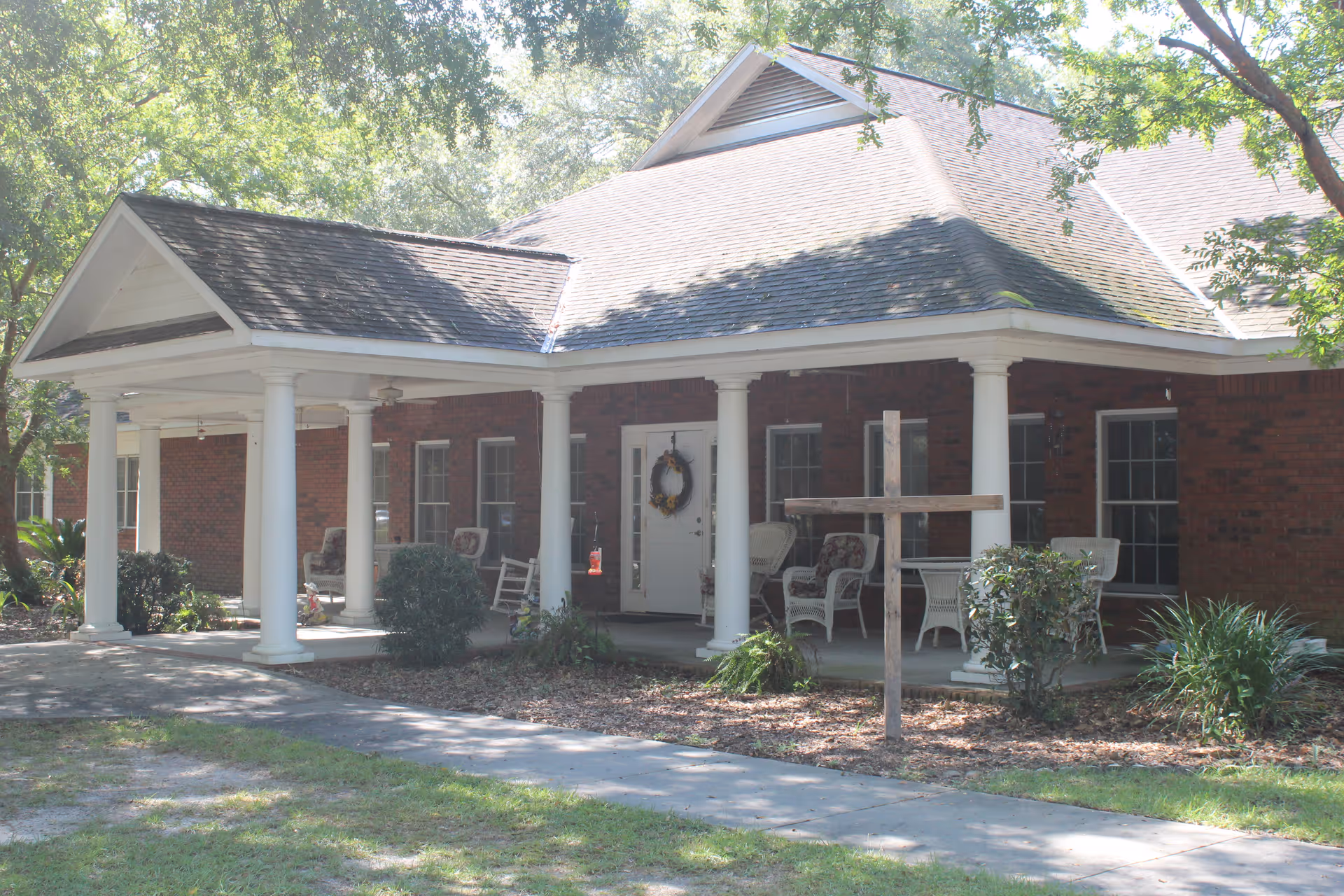 Front exterior view of a brick building with a covered porch supported by white columns. The porch has several chairs and a table, with a wreath hanging on the front door. There is a wooden cross planted in the ground near the walkway leading to the entrance, surrounded by bushes and trees.