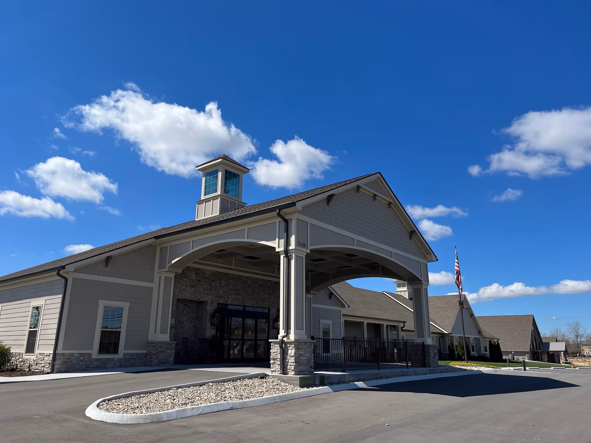 Front exterior of a beige senior living facility with a covered entrance/porte-cochere, stone accents, and an American flag under a blue sky.