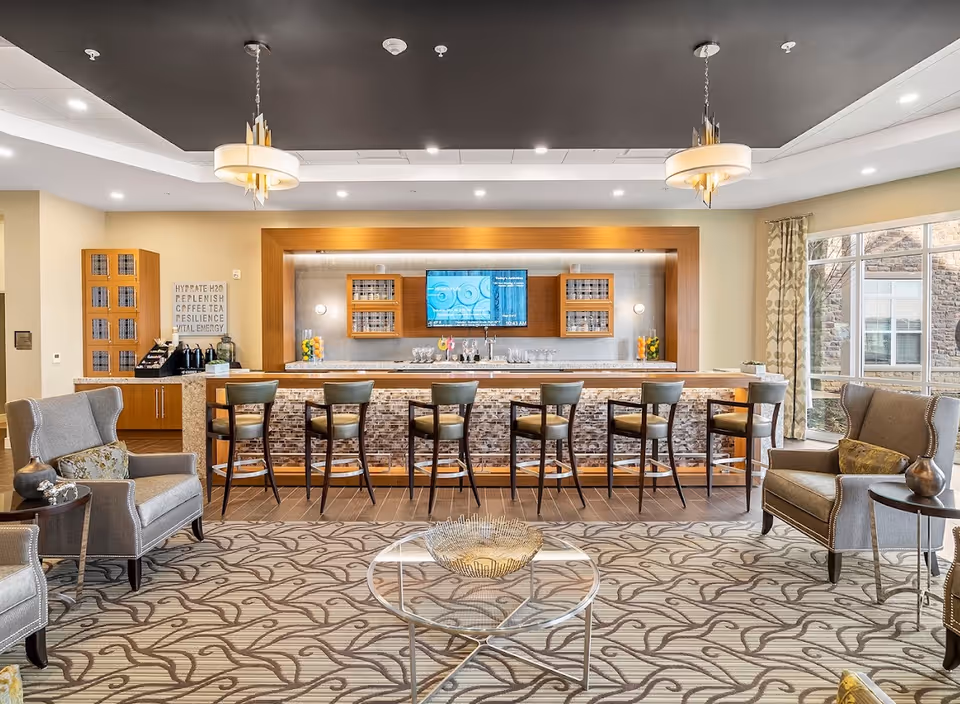 A modern lounge area with a bar counter featuring seven high chairs. Behind the bar is a wall-mounted TV flanked by wooden cabinets with glass doors. The room has a patterned carpet, two armchairs with side tables on either side of the bar, and large windows with patterned curtains letting in natural light.
