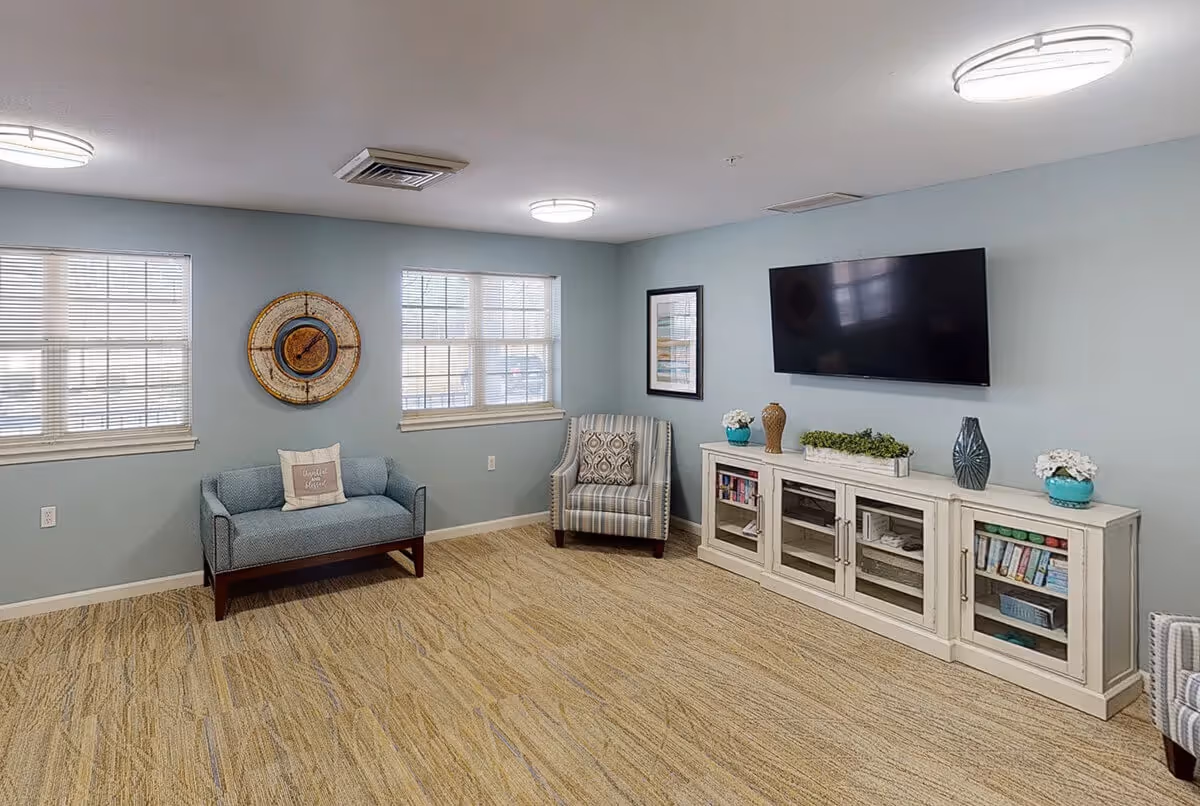 A cozy living room with light blue walls and beige carpet. The room features a small blue loveseat with a decorative pillow, a striped armchair, and a white media console with glass doors holding books and decorative items. A large flat-screen TV is mounted on the wall above the console. Two windows with blinds allow natural light into the room, and a round wall clock hangs between them. The ceiling has three modern circular light fixtures.