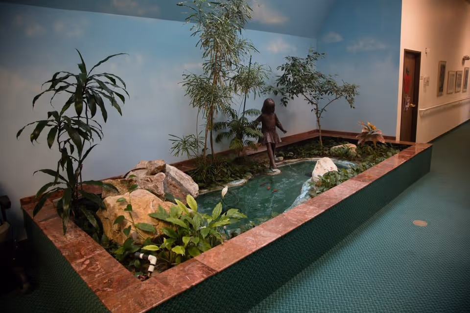 Indoor planted water feature with rocks, a small pond and a statue of a girl in a carpeted hallway.