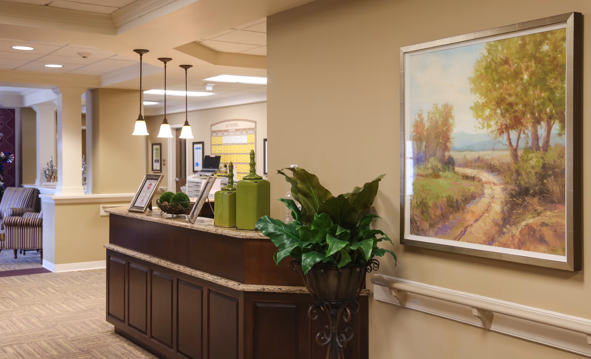 Reception area in a senior living facility with a dark wooden desk topped with decorative green vases and framed pictures. There are three pendant lights hanging above the desk. A large framed painting of a scenic landscape with trees and a path is on the wall to the right. In the background, there is a seating area with striped chairs and a bulletin board labeled 'Activities'.