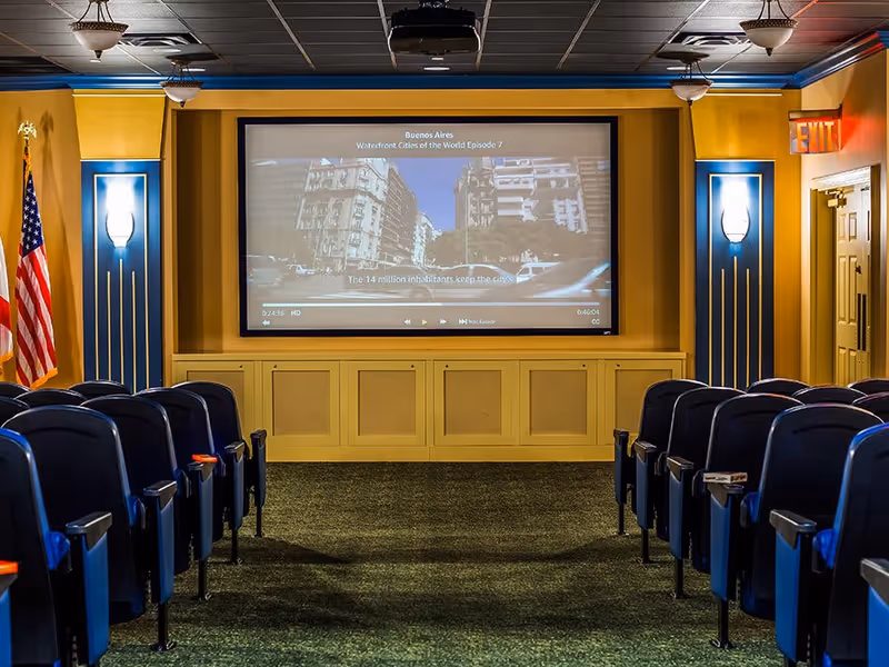 Interior view of a small theater or media room with rows of blue cushioned seats facing a large screen displaying a video about Buenos Aires. The room has yellow walls with blue accents, wall sconces, an American flag on the left, and an exit sign on the right.