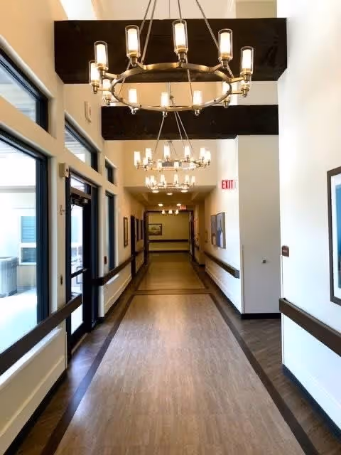 Well-lit interior hallway with hanging chandeliers, exposed wooden beams, wall handrails, framed artwork, and windows along one side.