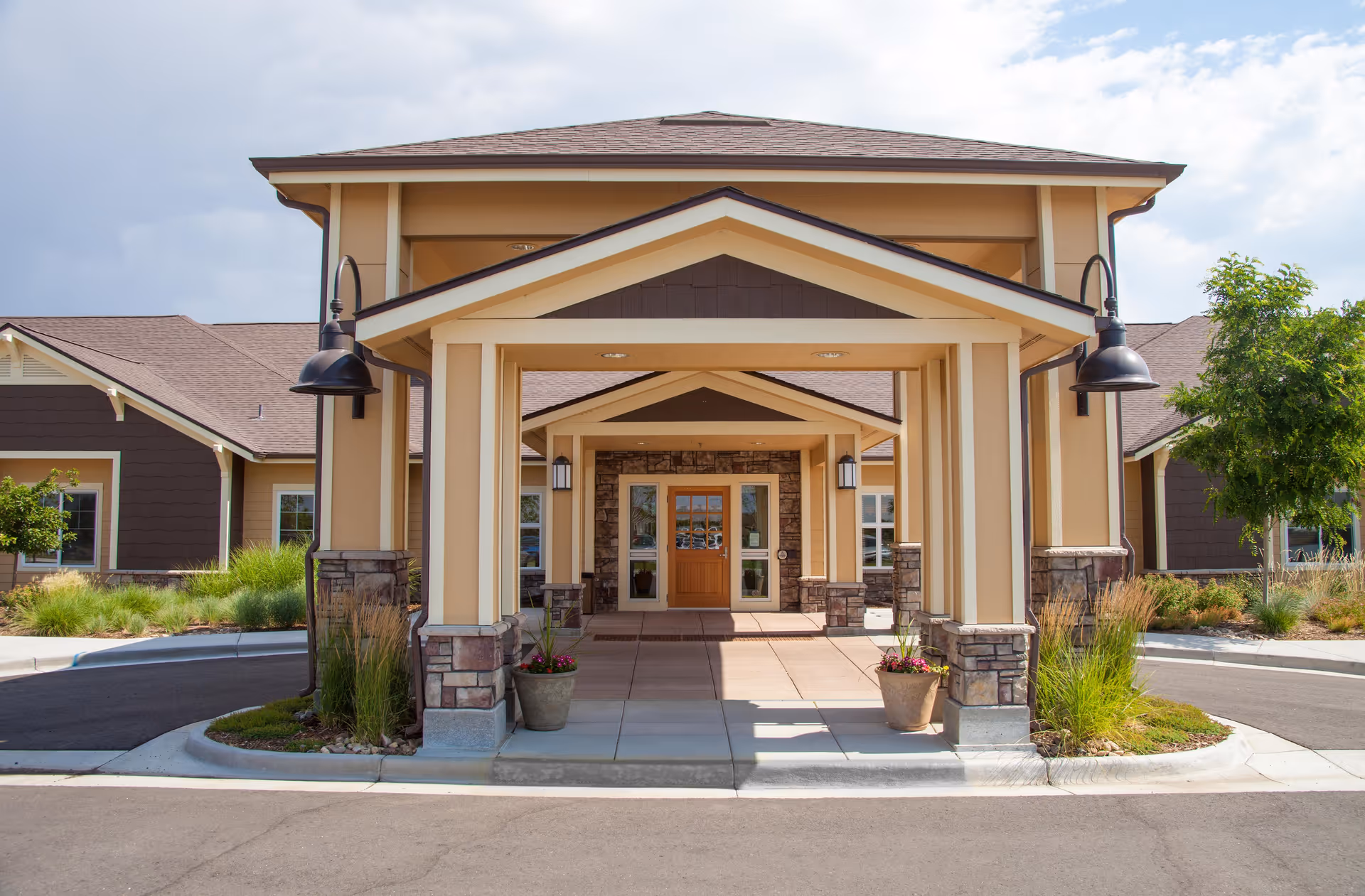 Front entrance of a single-story building with a covered walkway supported by columns, potted plants on either side, and outdoor lamps mounted on the columns. The building has a beige and brown exterior with a peaked roof and landscaping around the driveway.