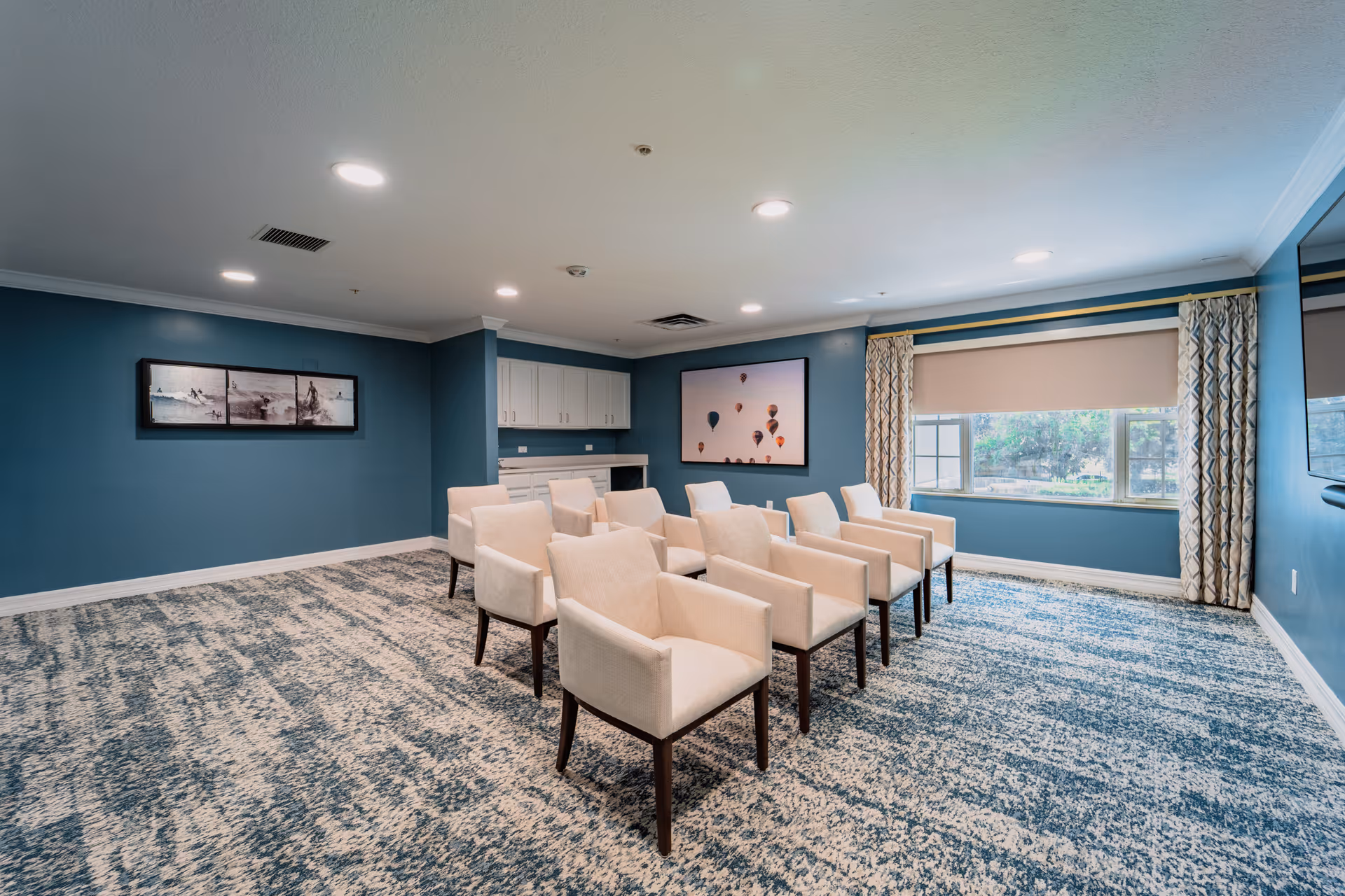 A room with blue walls and patterned carpet featuring two rows of beige upholstered chairs facing a large window with patterned curtains. The room has recessed ceiling lights, white cabinetry along one wall, and two framed pictures on the walls, one depicting hot air balloons and the other a black and white scene.