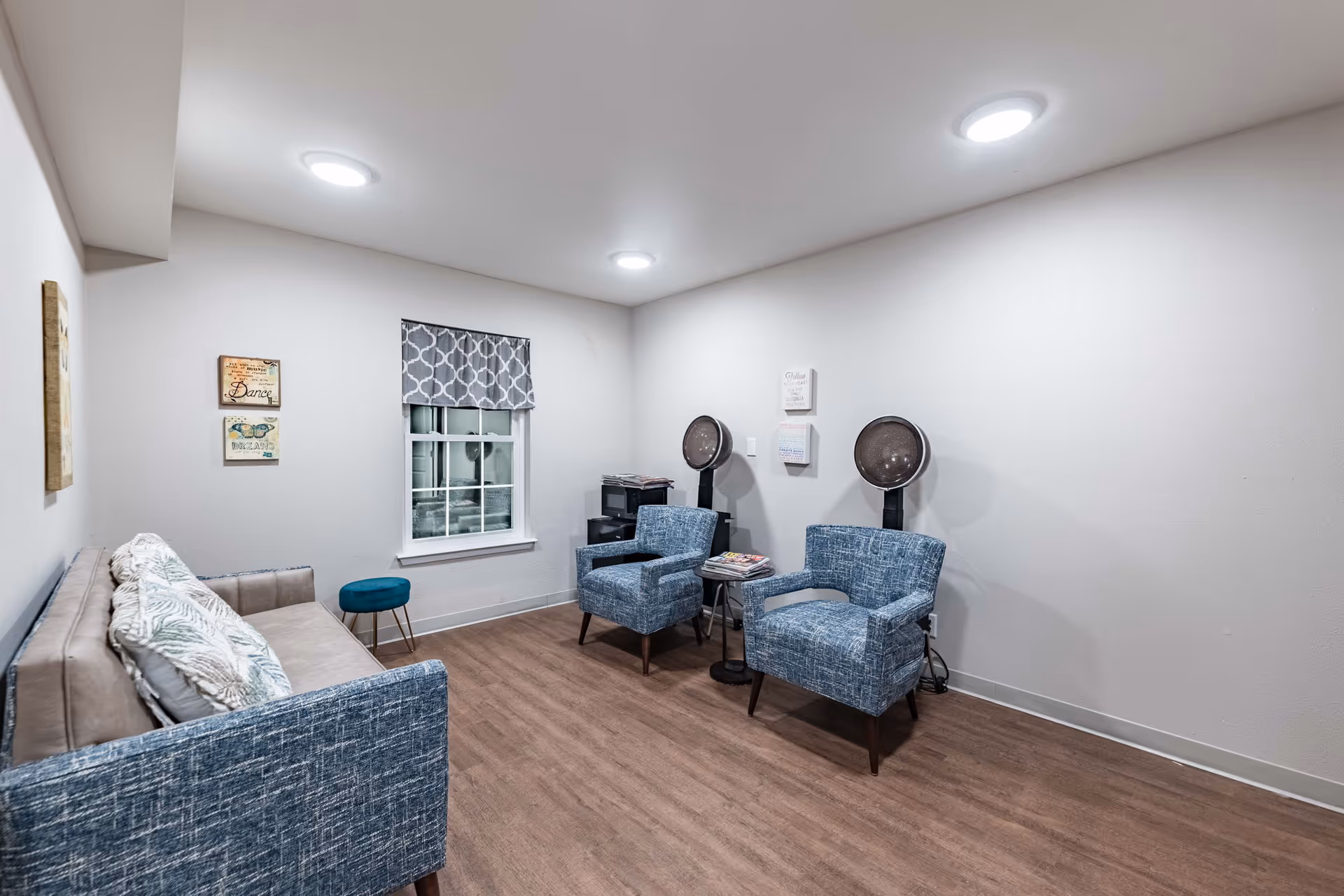 A cozy senior living room with a beige sofa adorned with patterned pillows, two blue armchairs, and a small teal stool. The room has wood flooring, white walls, a window with a patterned valance, and two hair drying stations with hooded dryers. Wall decorations include framed inspirational quotes.
