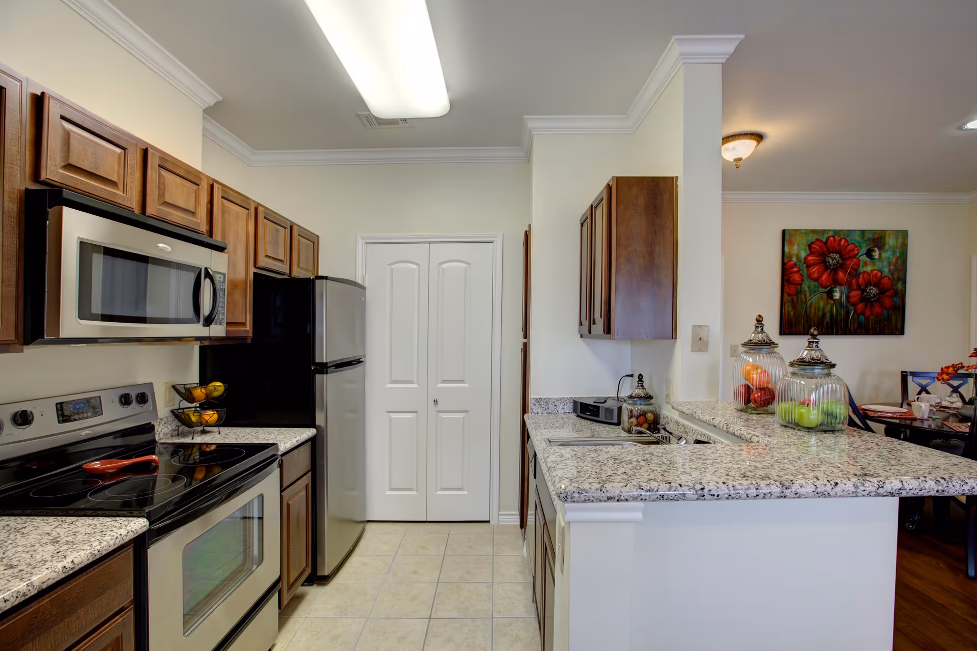 A modern kitchen with granite countertops, wooden cabinets, a stainless steel microwave, stove, and refrigerator. The kitchen has a tiled floor and a white double-door pantry at the end. On the right side, there is a breakfast bar with decorative glass jars containing fruit, and a glimpse of a dining area with a colorful floral painting on the wall.