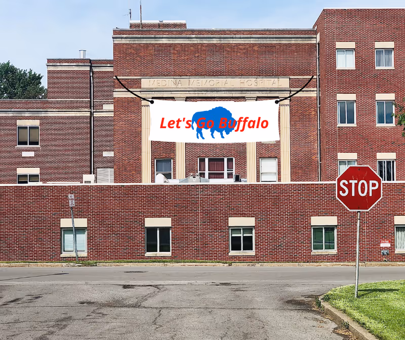 Front view of a red-brick hospital building with a large 'Let's Go Buffalo' banner hanging above and a stop sign in the foreground.