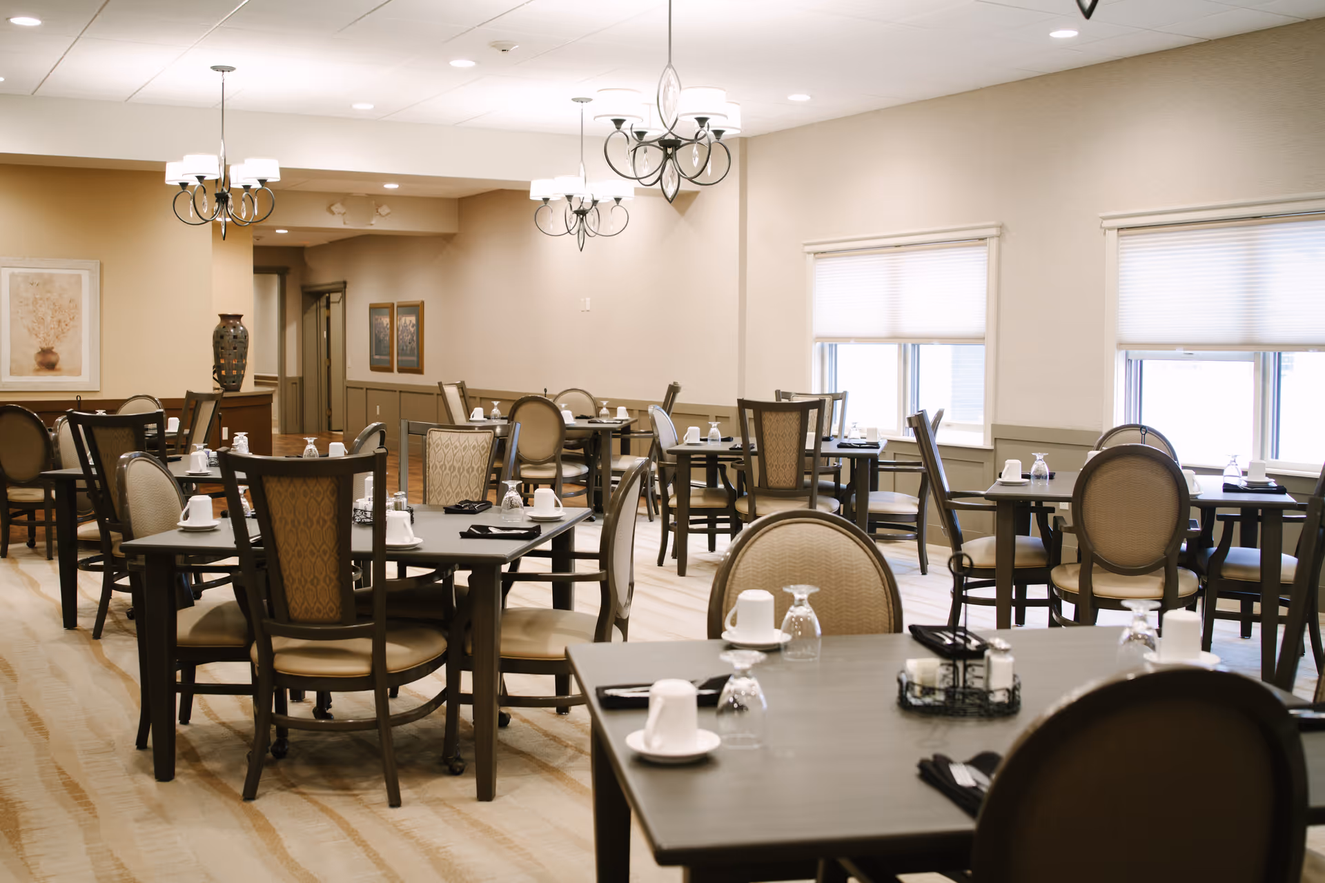 A well-lit dining room with multiple tables and chairs arranged neatly. Each table is set with white cups, upside-down glasses, black napkins, and silverware. The room has beige walls, large windows with blinds, and decorative light fixtures hanging from the ceiling. There are framed artworks and a large vase on a side table in the background.