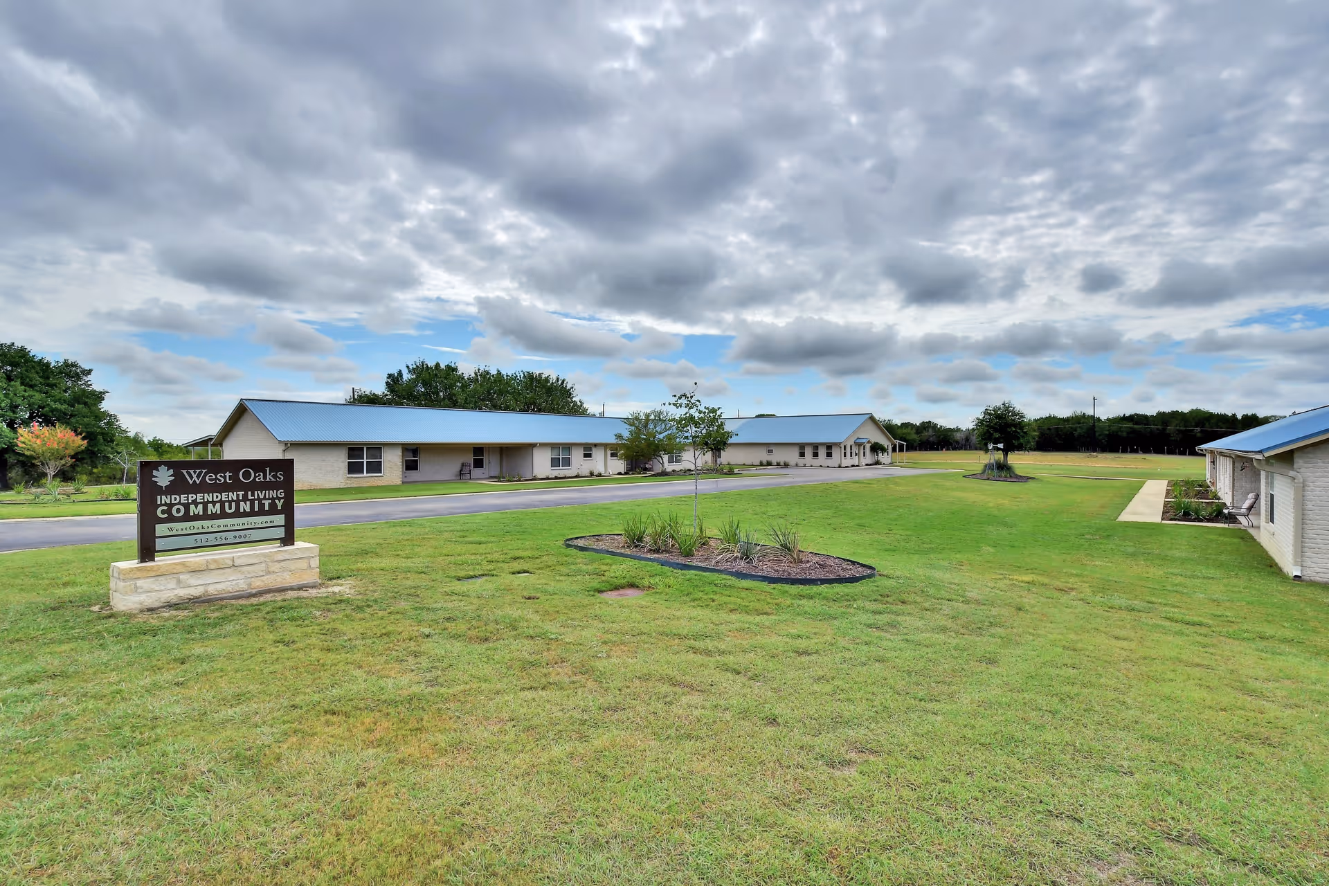 One-story beige buildings with blue metal roofs and a lawn, with a sign reading 'West Oaks Independent Living Community' under a cloudy sky.