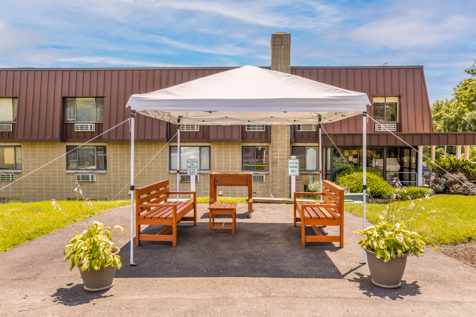 Outdoor seating area with two wooden benches, a small wooden table, and a foosball table under a white canopy tent. The area is paved and flanked by two potted plants with green foliage. In the background is a two-story building with brown siding and several windows, some with air conditioning units. The sky is partly cloudy.