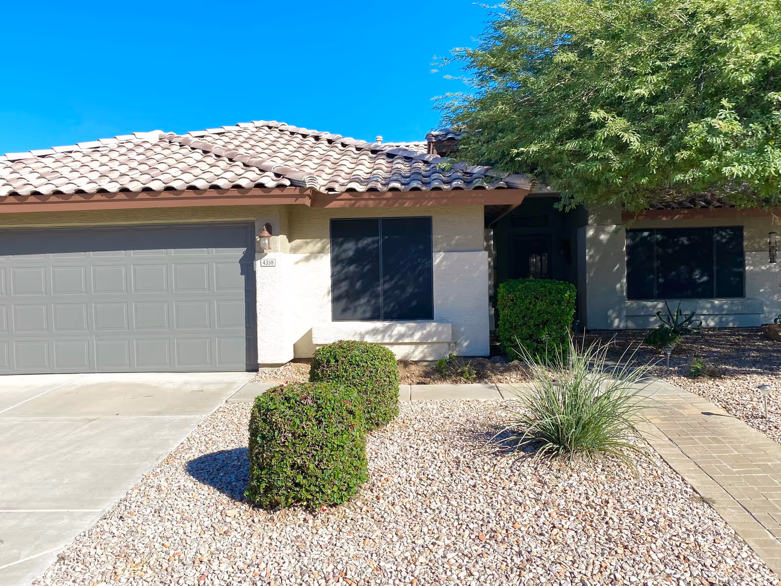 Single-story stucco house with a tiled roof, attached garage, gravel front yard, trimmed bushes, and a paved walkway to the entrance.